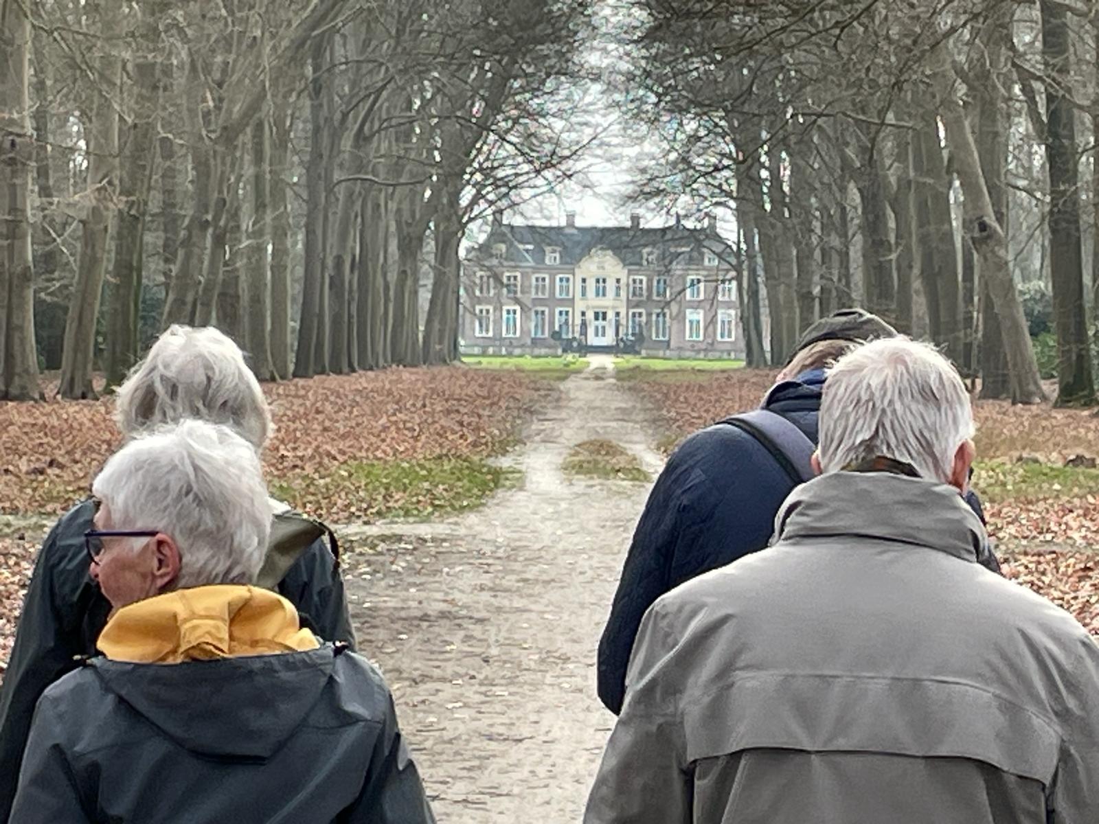 Vier mensen wandelen naar een statig herenhuis langs een bomenlaan in de herfst.