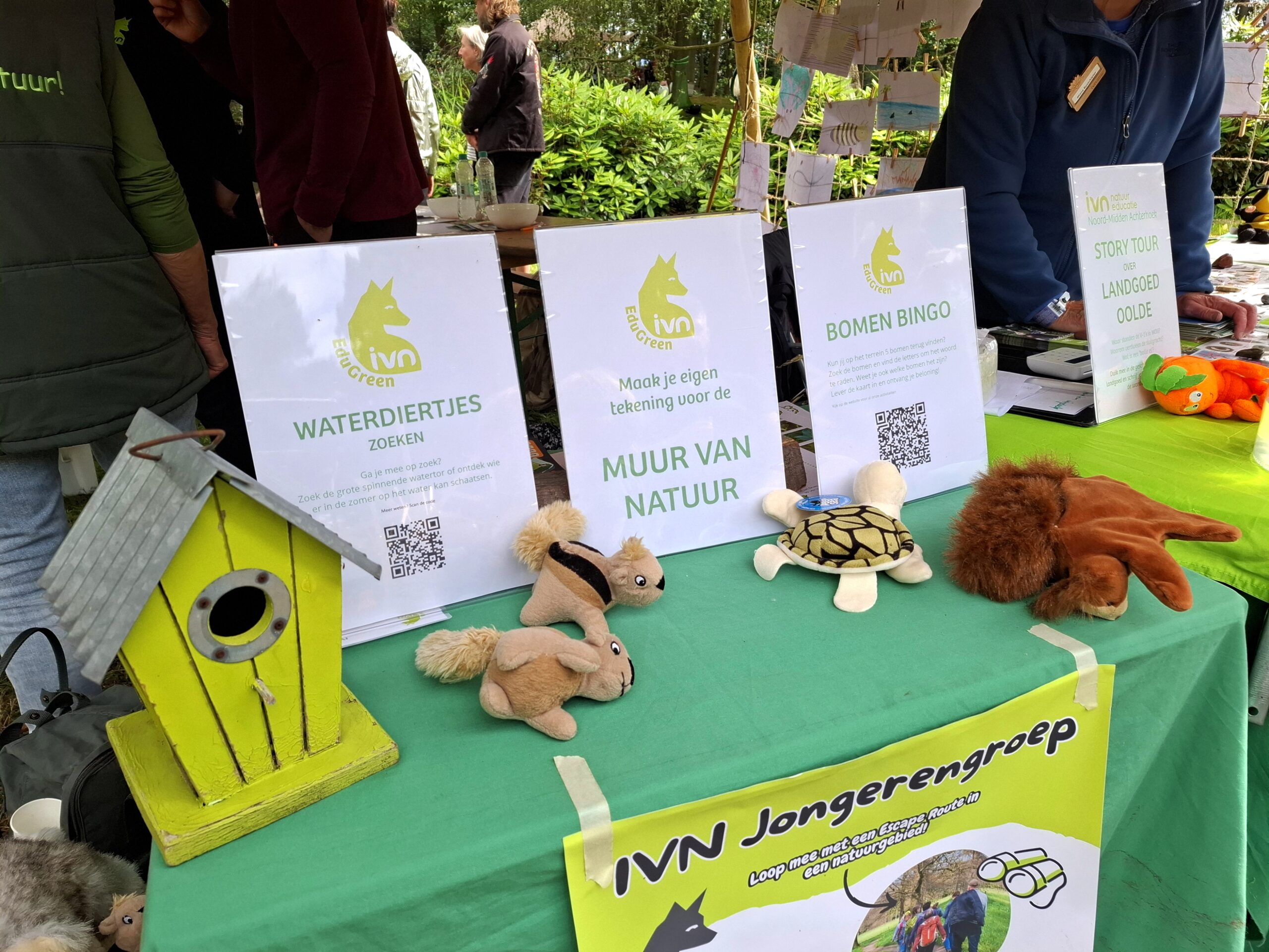 Informatietafel met knuffeldieren, vogelhuisje en IVN-activiteitposters in een groene omgeving.