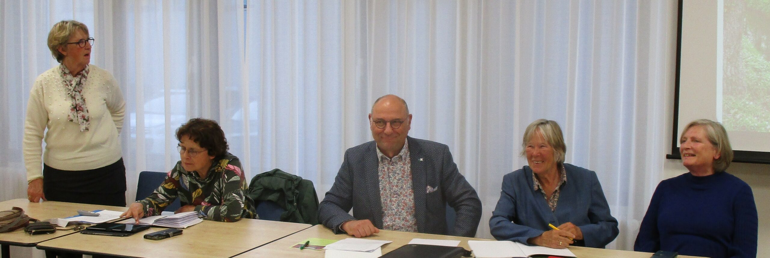 Vijf mensen zitten aan een tafel in een vergaderruimte met witte gordijnen.