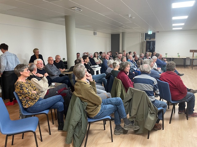 Een groep mensen zit in een zaal met blauwe stoelen, aandachtig luisterend naar een spreker.