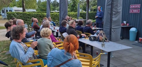 Groep mensen luistert naar een spreker bij een buitenbijeenkomst met tafels en gele stoelen.