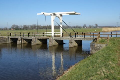 Houten ophaalbrug over kanaal in groen landschap onder blauwe lucht.