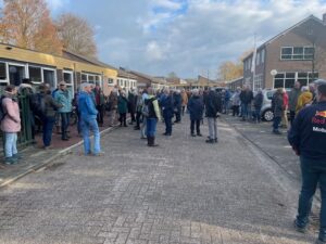Groep mensen verzameld buiten een gebouw op een bewolkte dag. Herfstbladeren op de achtergrond.