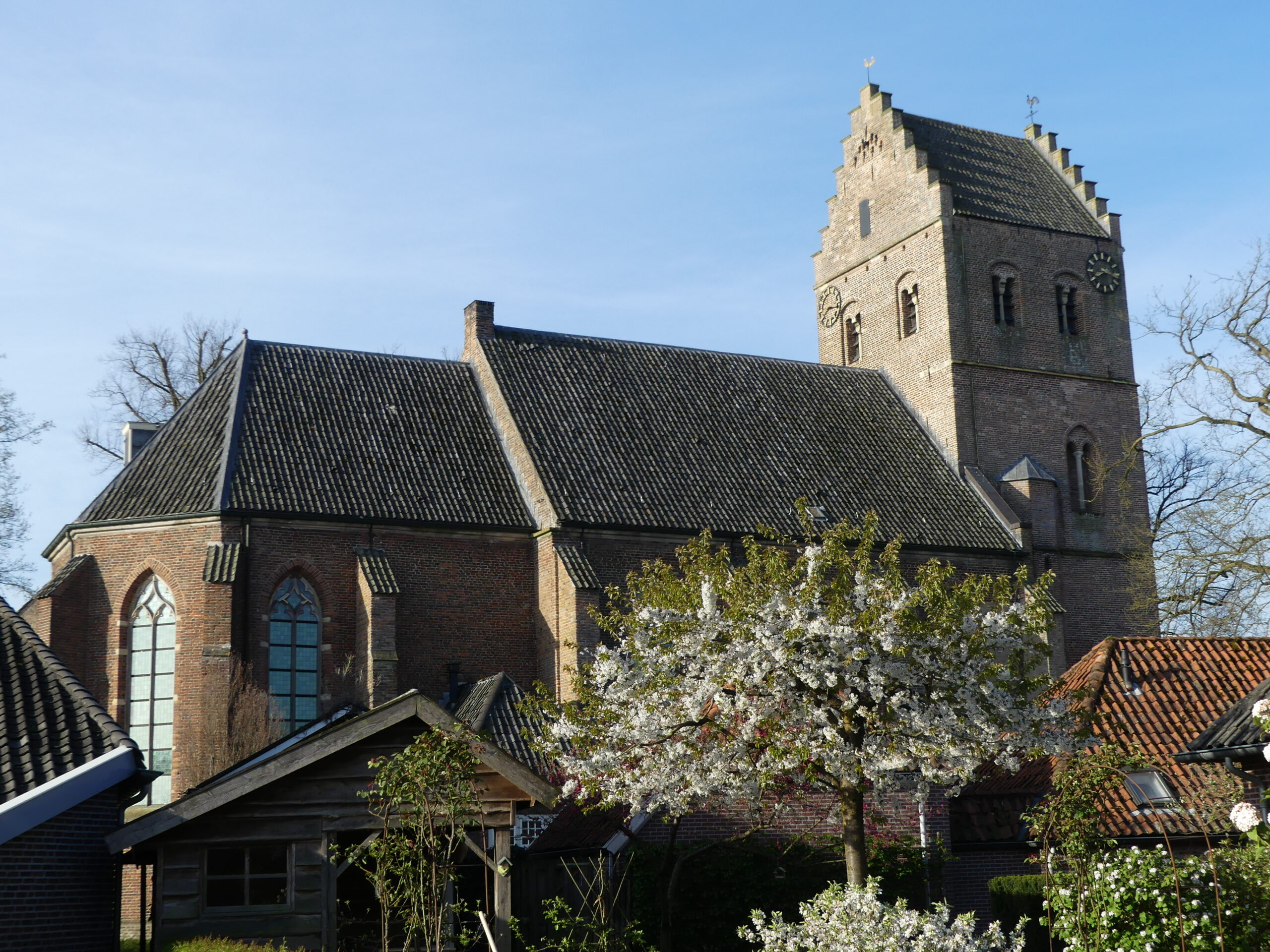 Kerk met bakstenen muren, kloktoren en bloeiende boom op de voorgrond onder een blauwe lucht.