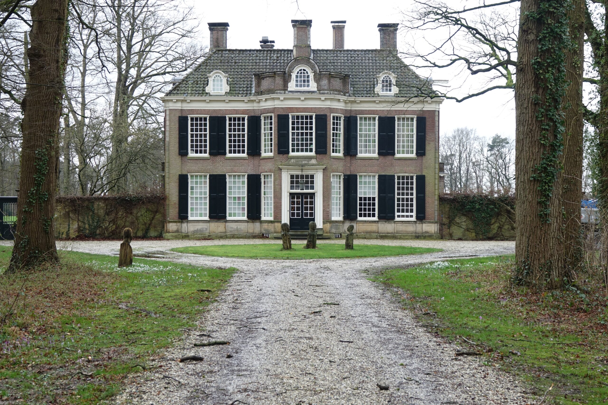 Historisch herenhuis met symmetrische gevel, omringd door bomen en een grindpad aan de voorkant.