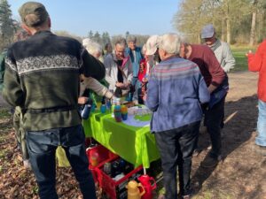 Groep mensen verzameld rond een tafel met thee en koffie in een parkachtige omgeving.