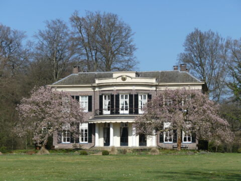 Groot landhuis met bloeiende bomen aan de voorzijde, omgeven door een groene tuin.