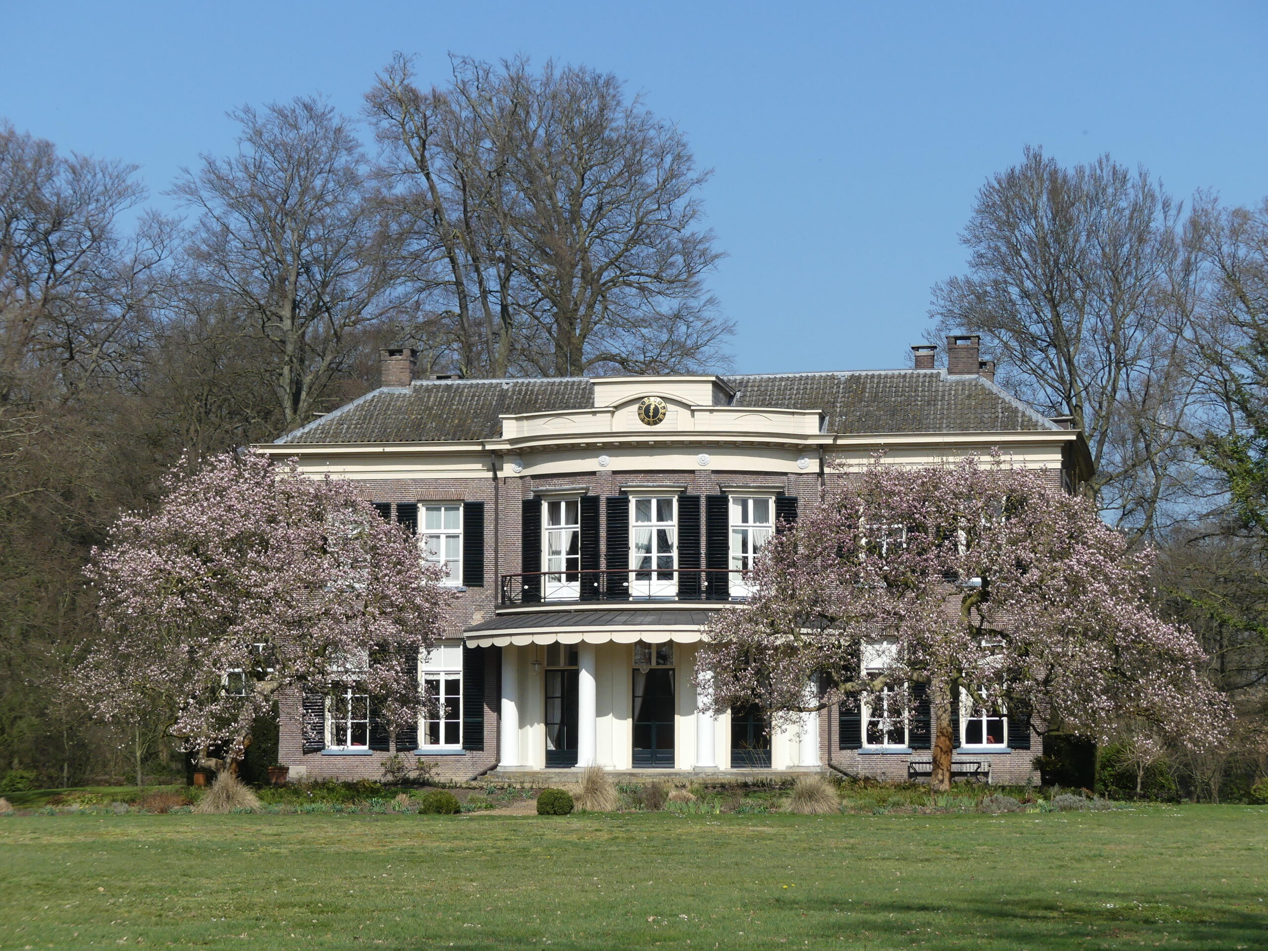 Groot landhuis met bloeiende bomen aan de voorzijde, omgeven door een groene tuin.