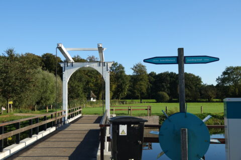 Houten brug met borden, omgeven door groen veld en bomen onder een heldere blauwe lucht.