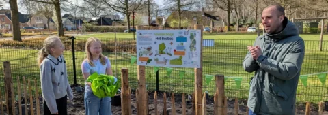 Twee kinderen en een man bij een bord van een nieuw voedselbos, in een groene buitenomgeving.