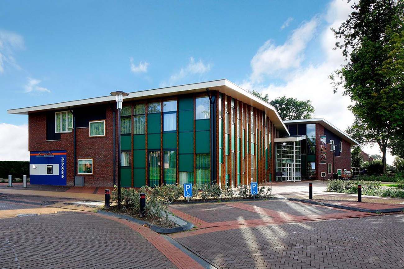 Modern bakstenen gebouw met groene panelen en Rabobank logo, omringd door bomen en parkeerplaatsen.