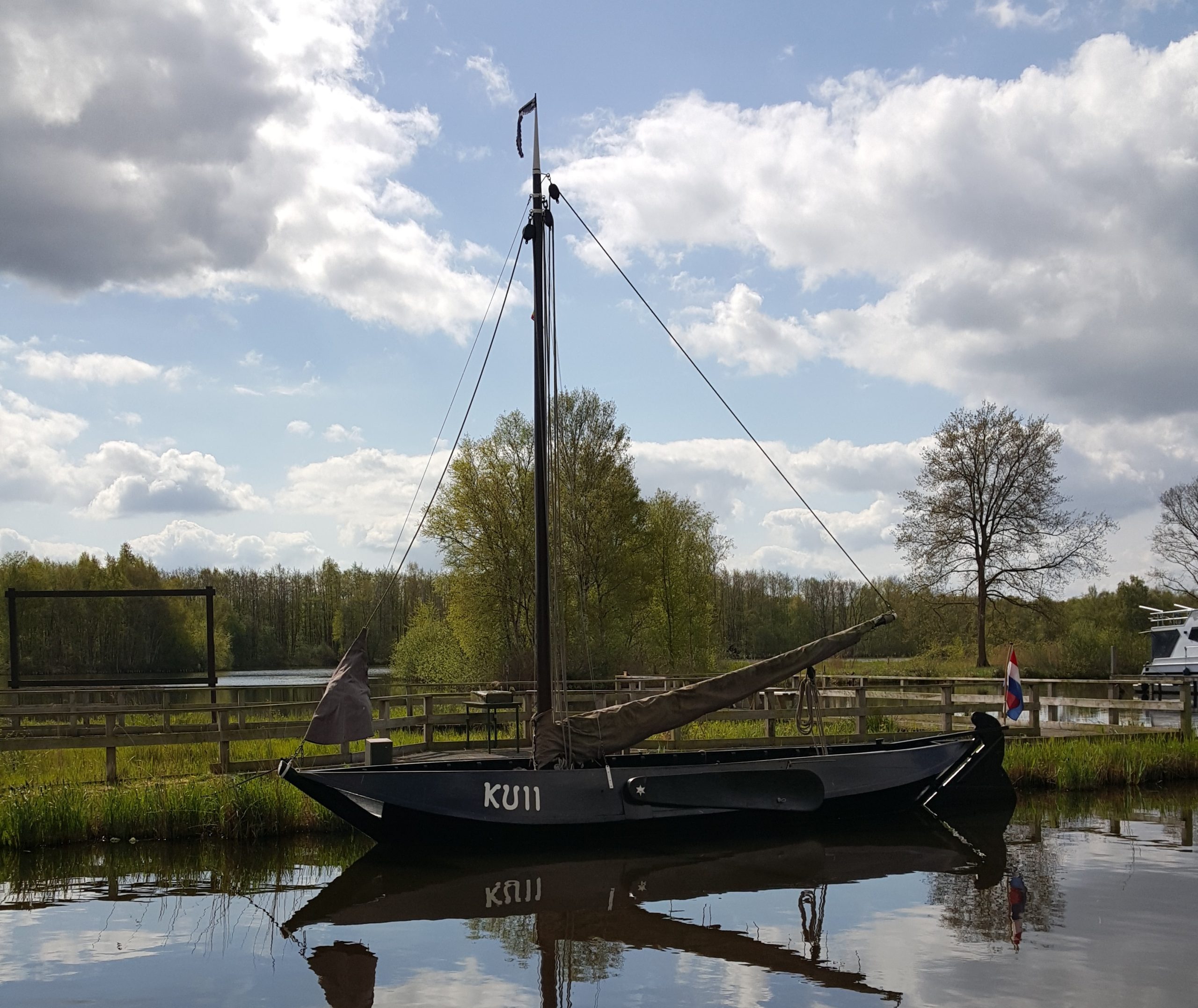 Historische boot met mast en Nederlandse vlag, dobbert op een rustige rivier, omgeven door bomen.