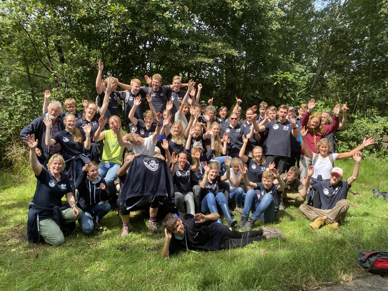 Een groep mensen, zwaaiend en lachend, poseert buiten in een groene, bosrijke omgeving.