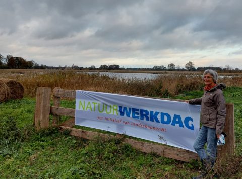 Vrouw bij een bord met "Natuurwerkdag" in een open, landelijk landschap met water en begroeiing.