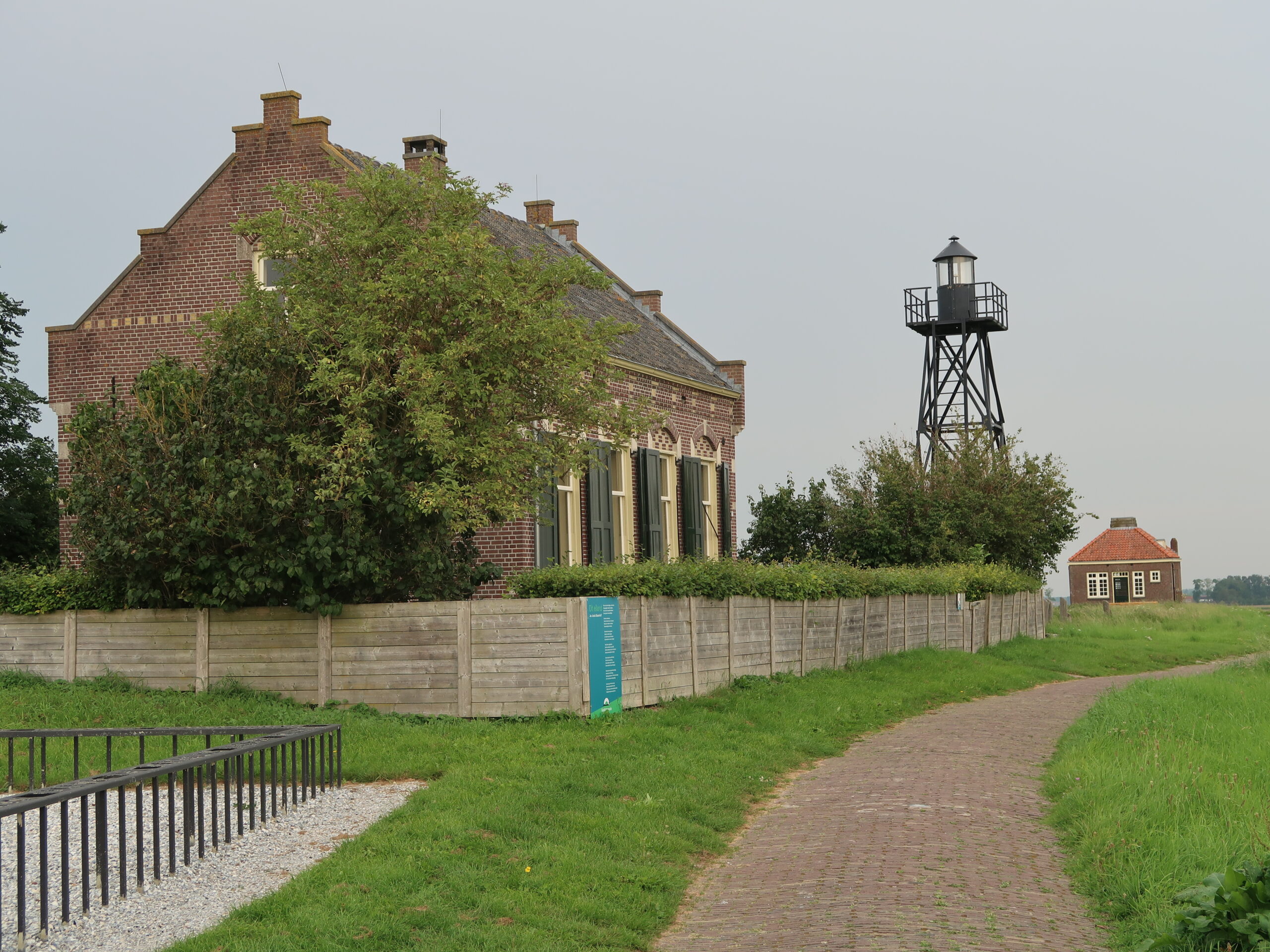 Bakstenen huis met groene luiken, hek, pad, vuurtoren en klein gebouw in een groene omgeving.