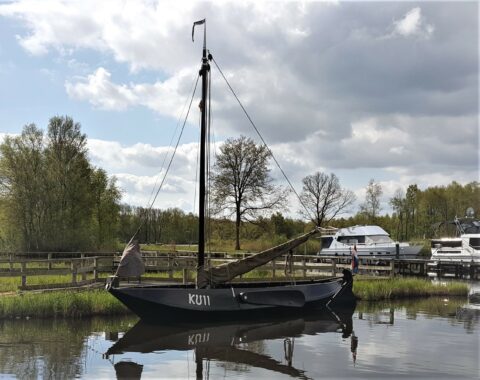 Traditionele boot met de naam KU11 aangemeerd bij een steiger. Wolkenlucht en groene bomen op achtergrond.