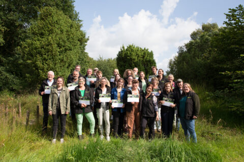 Groep mensen buiten in groene omgeving, poseren met certificaten onder blauwe lucht.