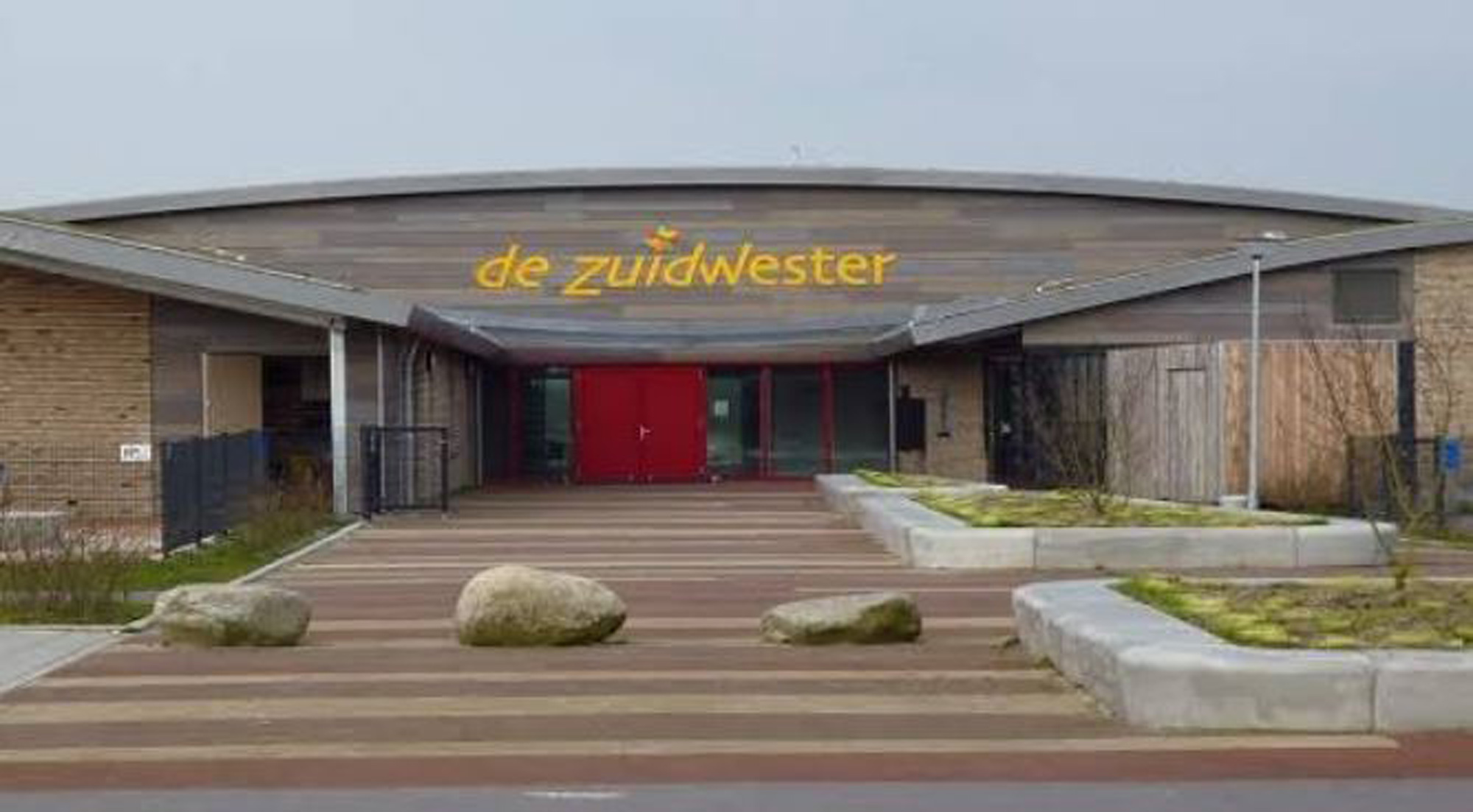Vooraanzicht van gebouw met rode deuren en gele tekst "de zuidwester" op houten gevel, stenen pad.