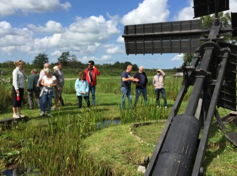 Groep mensen bij een windmolen in een groen polderlandschap onder een blauwe lucht met wolken.