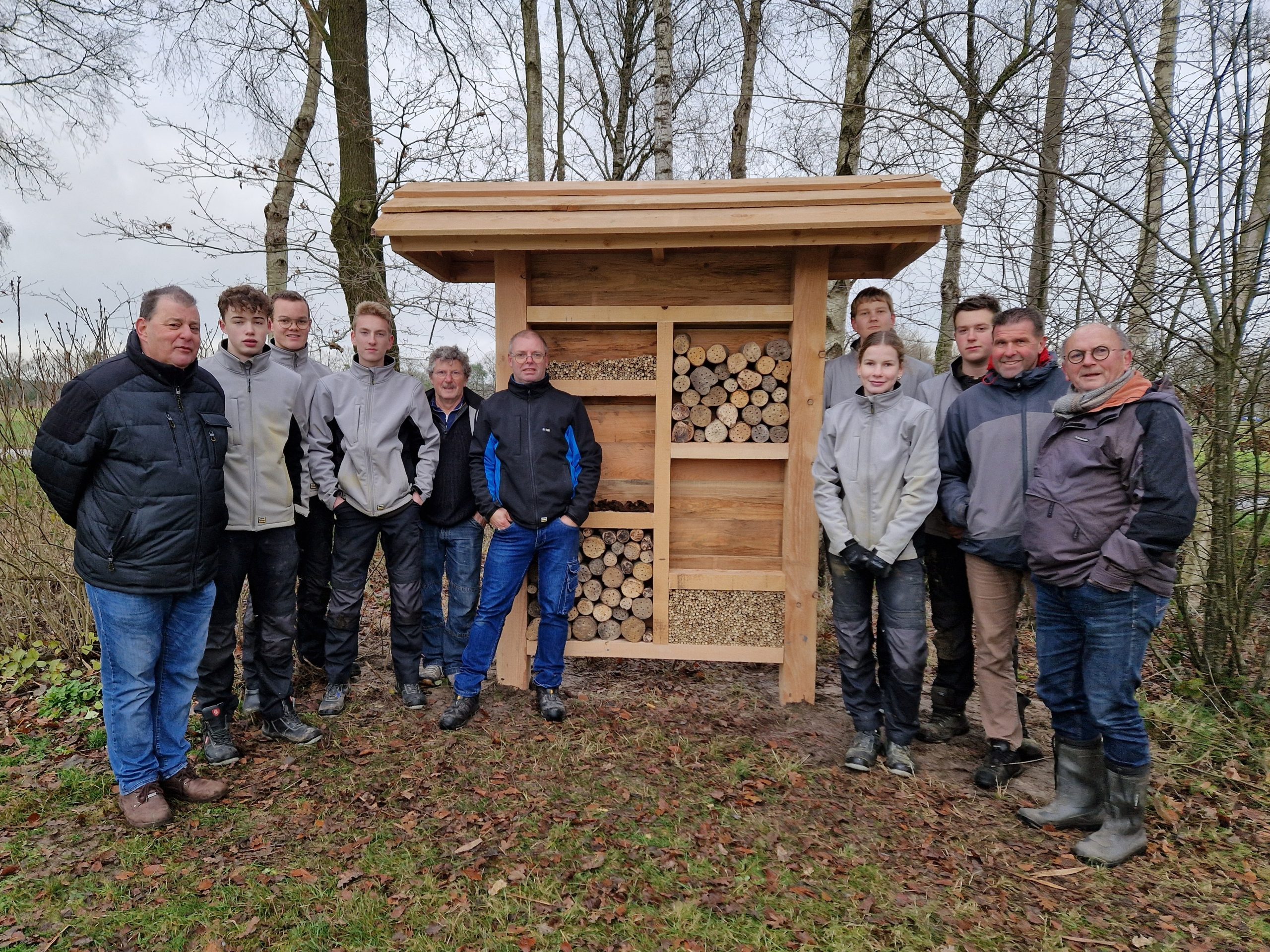Groep mensen poseert naast een insectenhotel in een bosrijke omgeving.