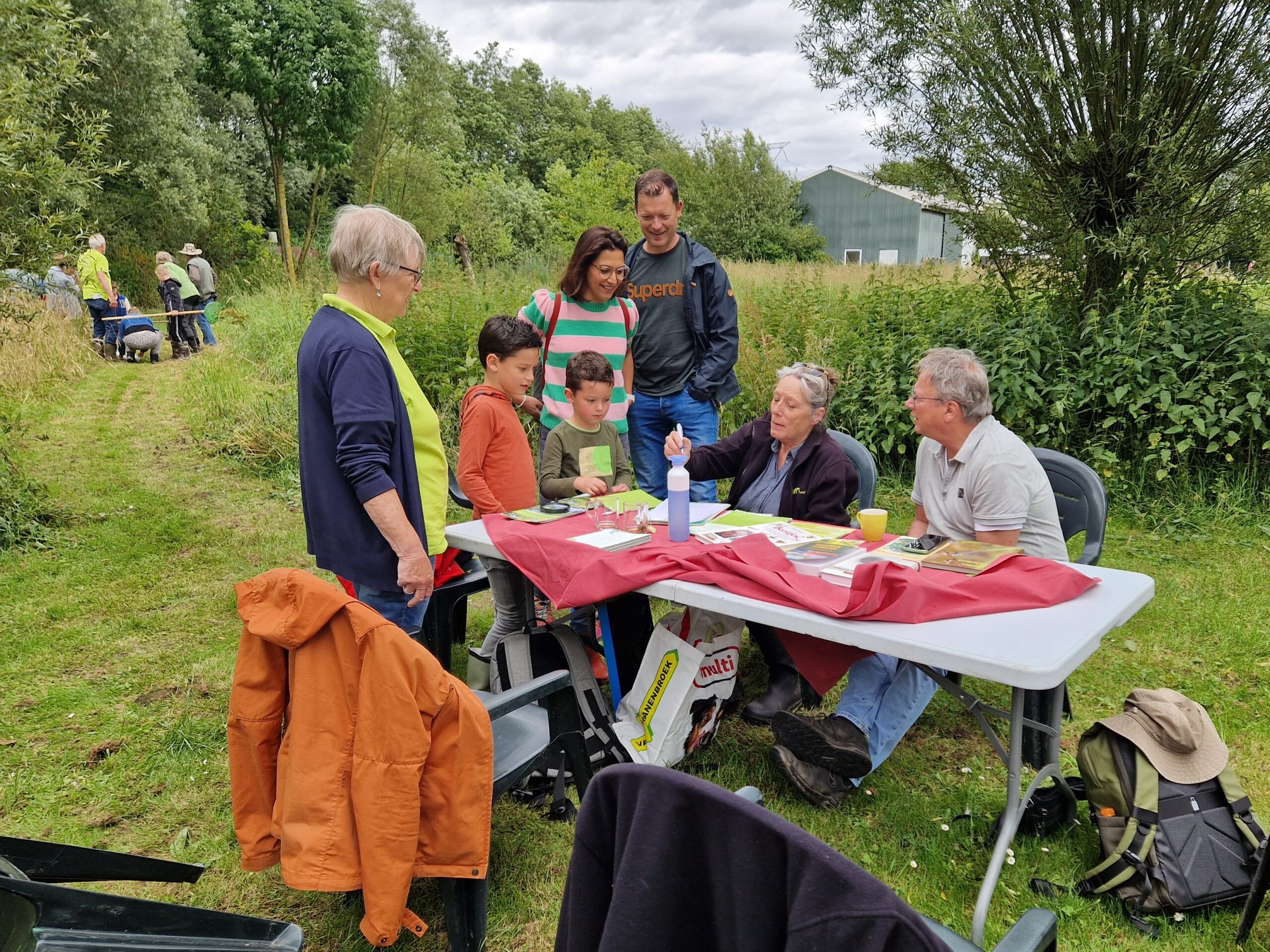 Groep mensen rond een tafel buiten, kinderen krijgen uitleg, andere mensen werken in de natuur.