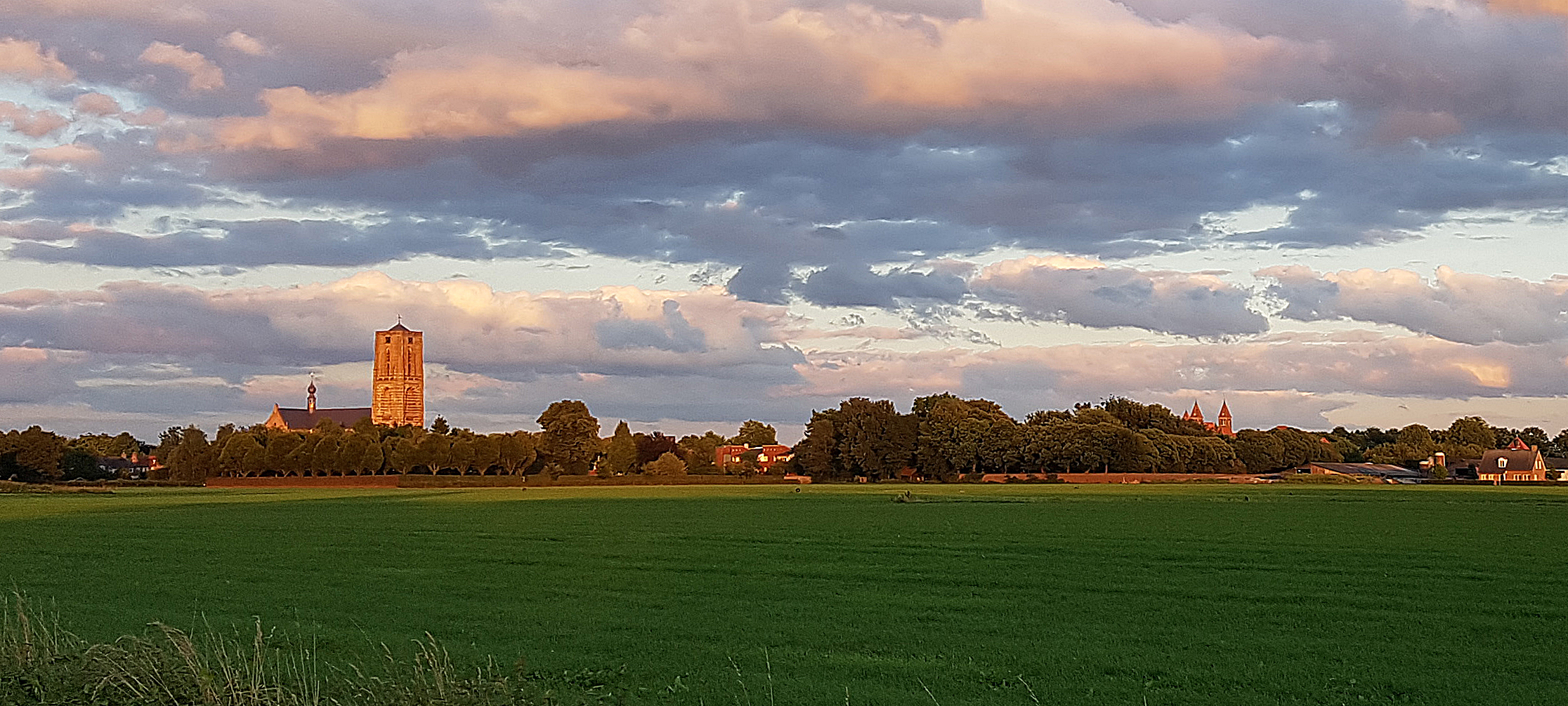 Landschap met kerktoren en dorp op de achtergrond, groene weilanden en kleurrijke wolkenluchten.