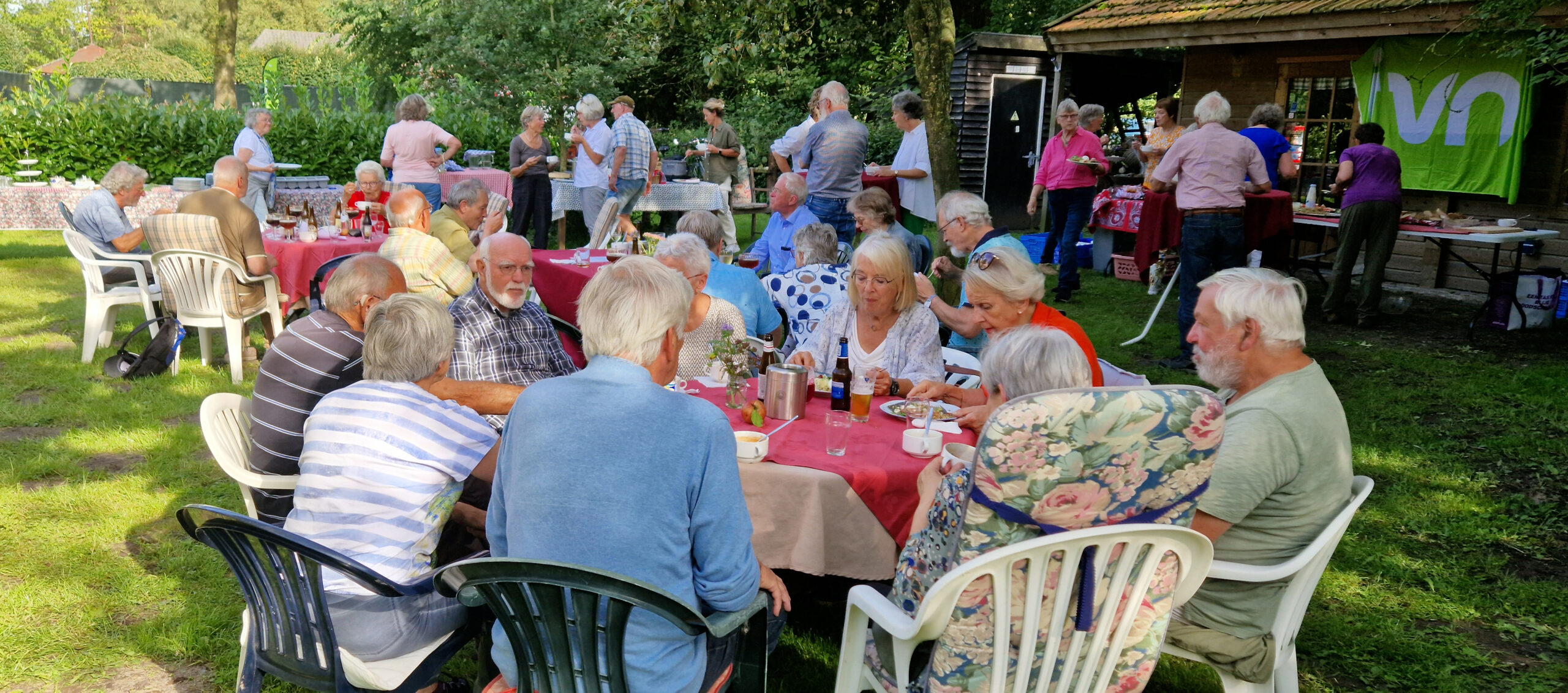 Groepsbijeenkomst in tuin, mensen zitten aan tafels, er is eten en drinken.