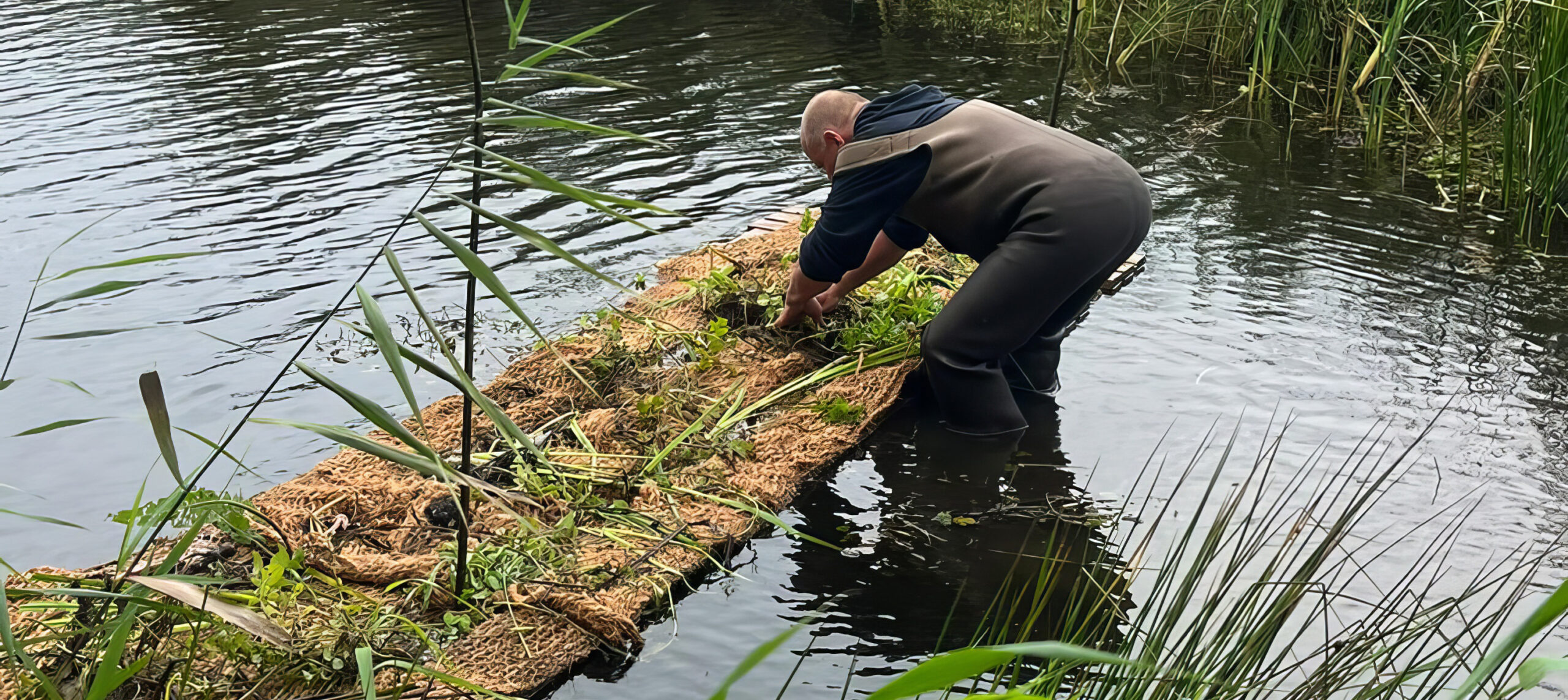 Persoon plant vegetatie op een drijvend eiland in een waterlichaam, omgeven door riet.
