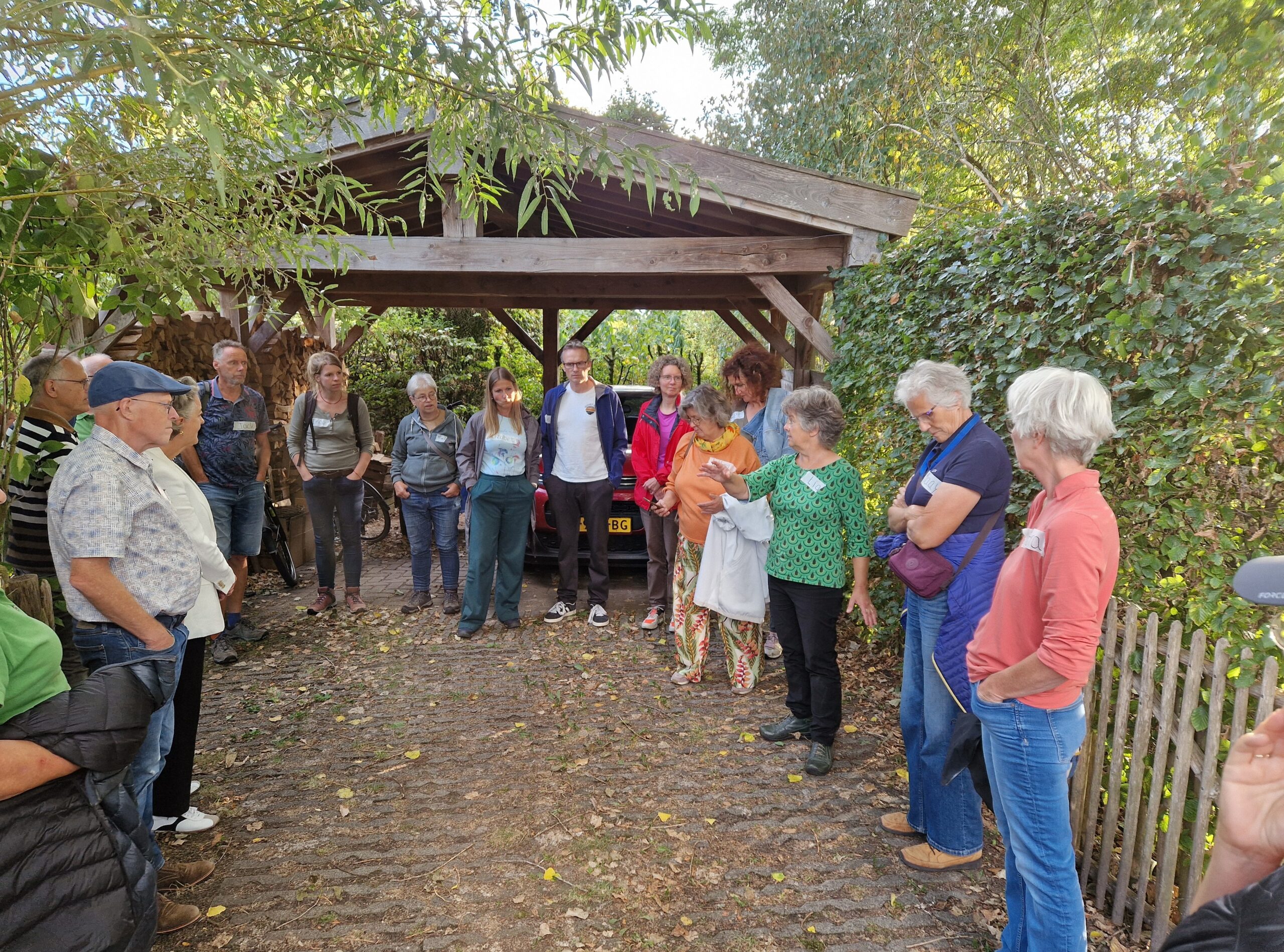 Een groep mensen staat in een kring onder een houten afdak in een groen, bosrijk gebied.