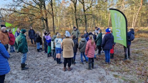 Groep mensen verzameld in een bos, met een groene vlag met logo.