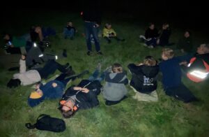 Groep mensen zit en ligt in het gras in het donker, buiten, mogelijk tijdens een nachtactiviteit.