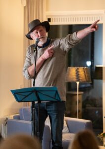 Man met hoed spreekt in microfoon en wijst naar rechts, voorleesstandaard met blauwe bladen.