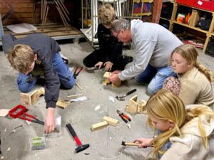 Kinderen en een volwassene timmeren samen houten objecten op de vloer van een werkplaats.