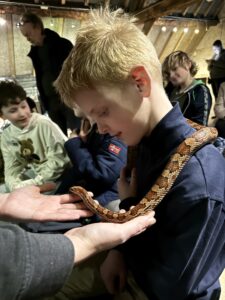 Jongen met blonde haren houdt een slang op zijn schouders, omringd door andere kinderen in een houten ruimte.
