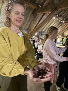 Vrouw in gele trui houdt een grote spin vast in een educatieve setting met kinderen op de achtergrond.