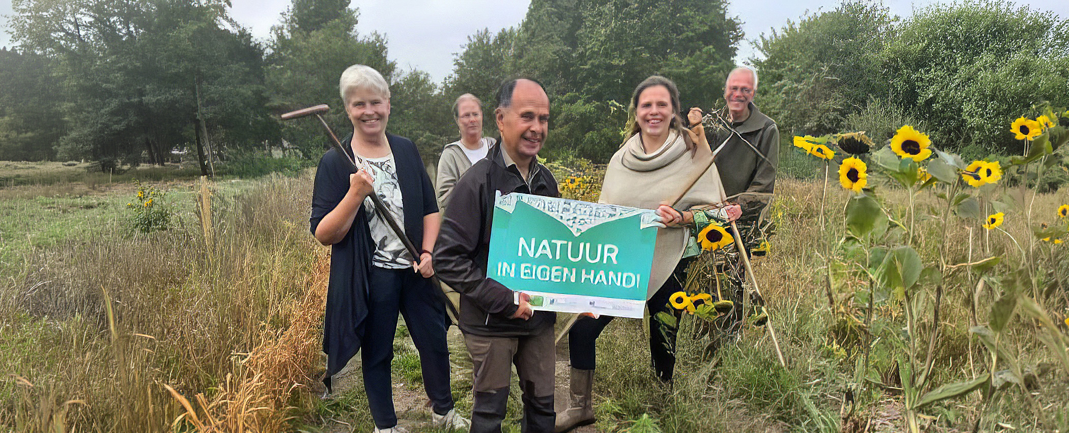 Groep mensen buiten met gereedschap en een bord "Natuur in eigen hand" bij zonnebloemen.