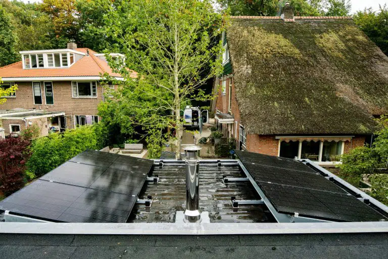 Dak met zonnepanelen, schoorsteen in het midden, omliggende huizen met bomen in de achtergrond.
