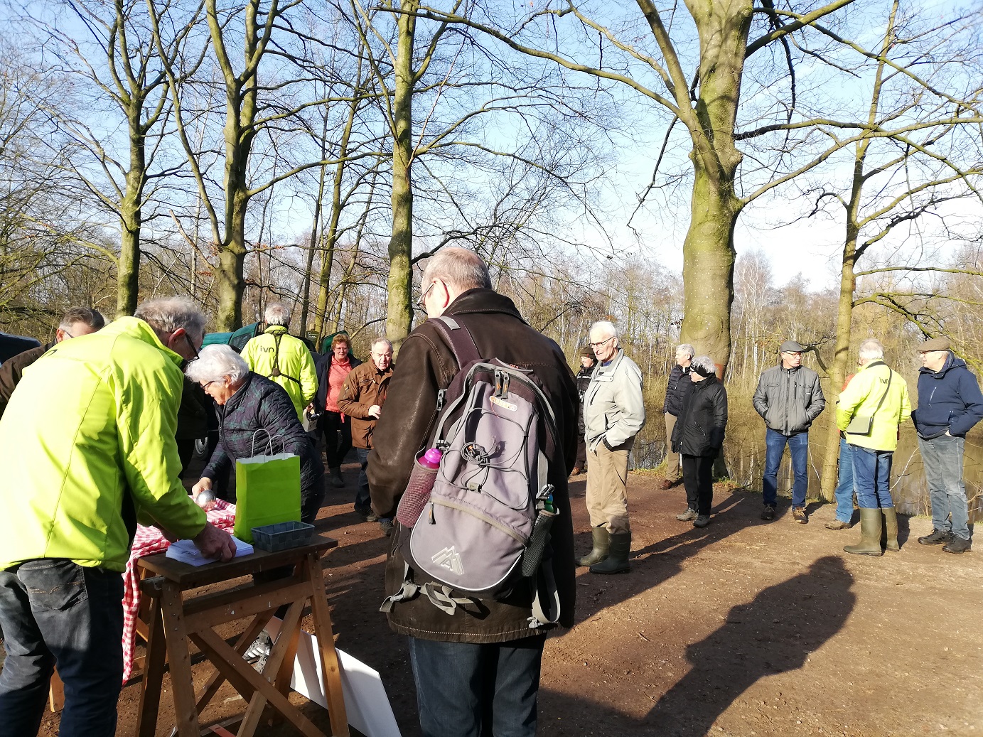 Groep mensen in park bij tafel met rood-wit kleed, sommigen dragen felgekleurde jasjes.