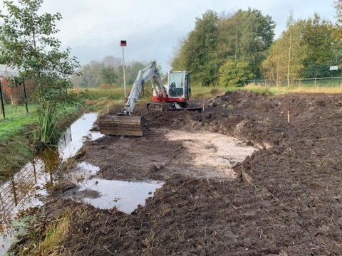 Graafmachine bij een modderige bouwplaats naast een smalle sloot, omringd door bomen.
