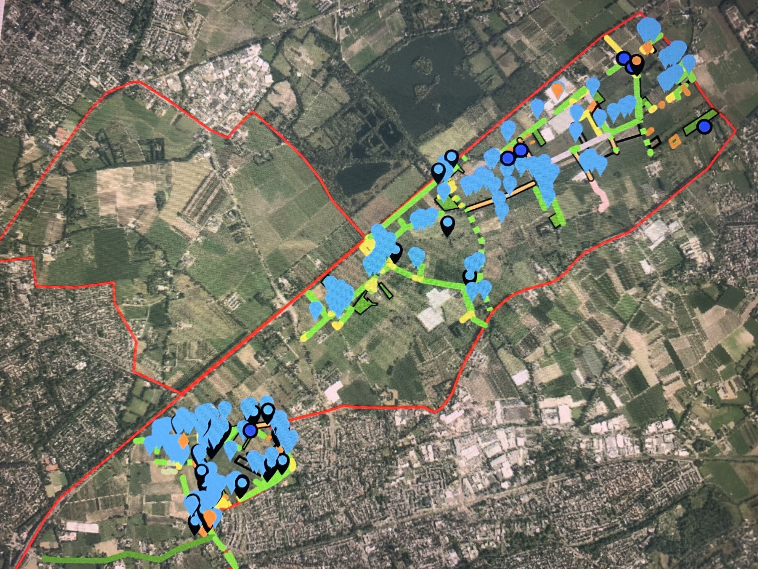 Luchtfoto met rode grenslijn, blauwe markeringen, groene en gele paden door landelijk gebied.