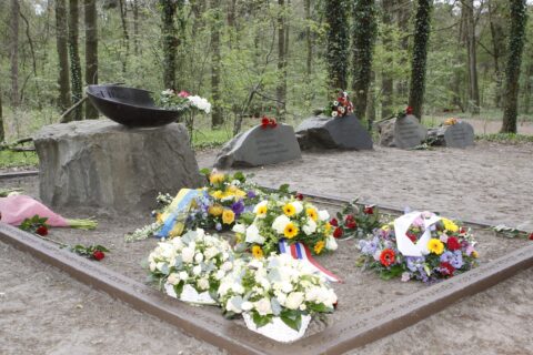 Gedenkplaats met bloemenkransen op stenen; bomen op achtergrond.