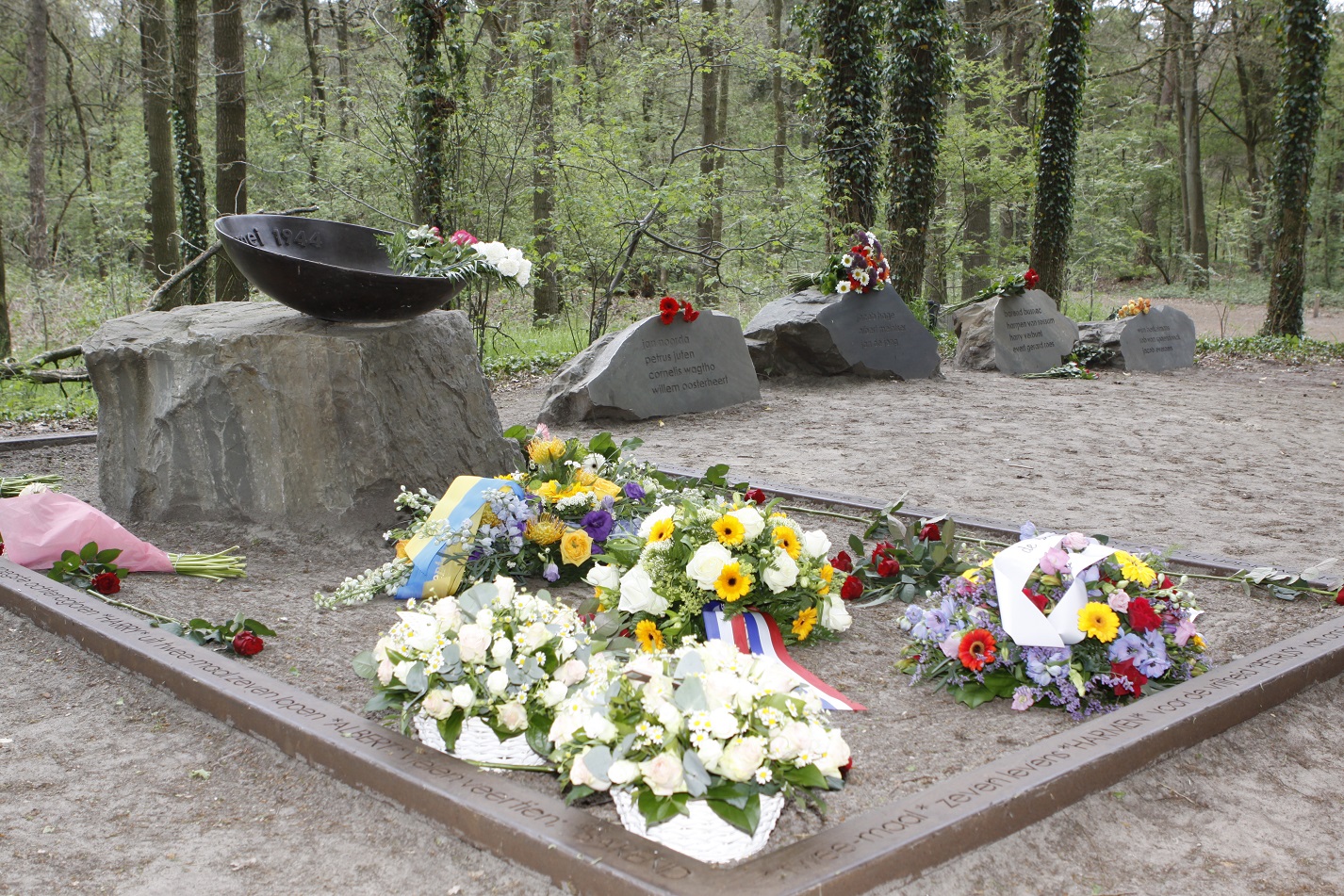 Gedenkplaats met bloemenkransen op stenen; bomen op achtergrond.