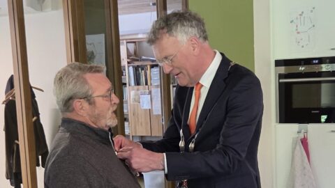 Een man in een pak speldt een lintje op bij een andere man in een huiselijke omgeving.