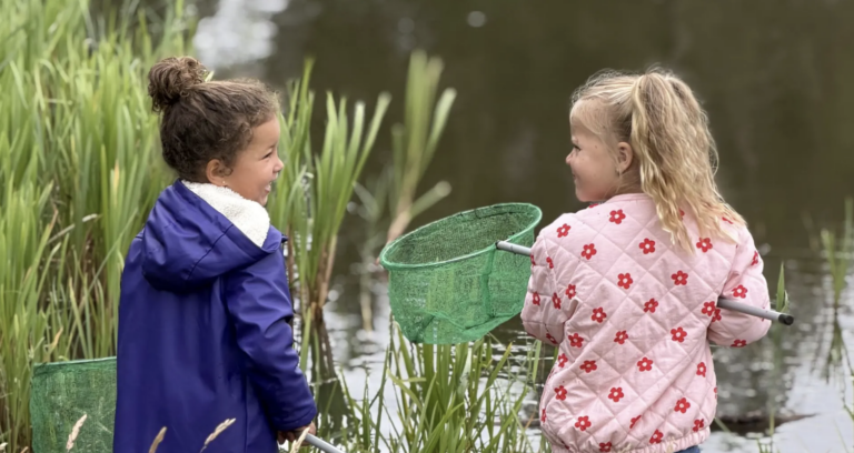 Twee kinderen met schepnetten bij een vijver, omringd door riet.
