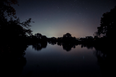 Nachtelijke hemel met sterren boven een stille rivier, omlijst door donkere silhouetten van bomen.