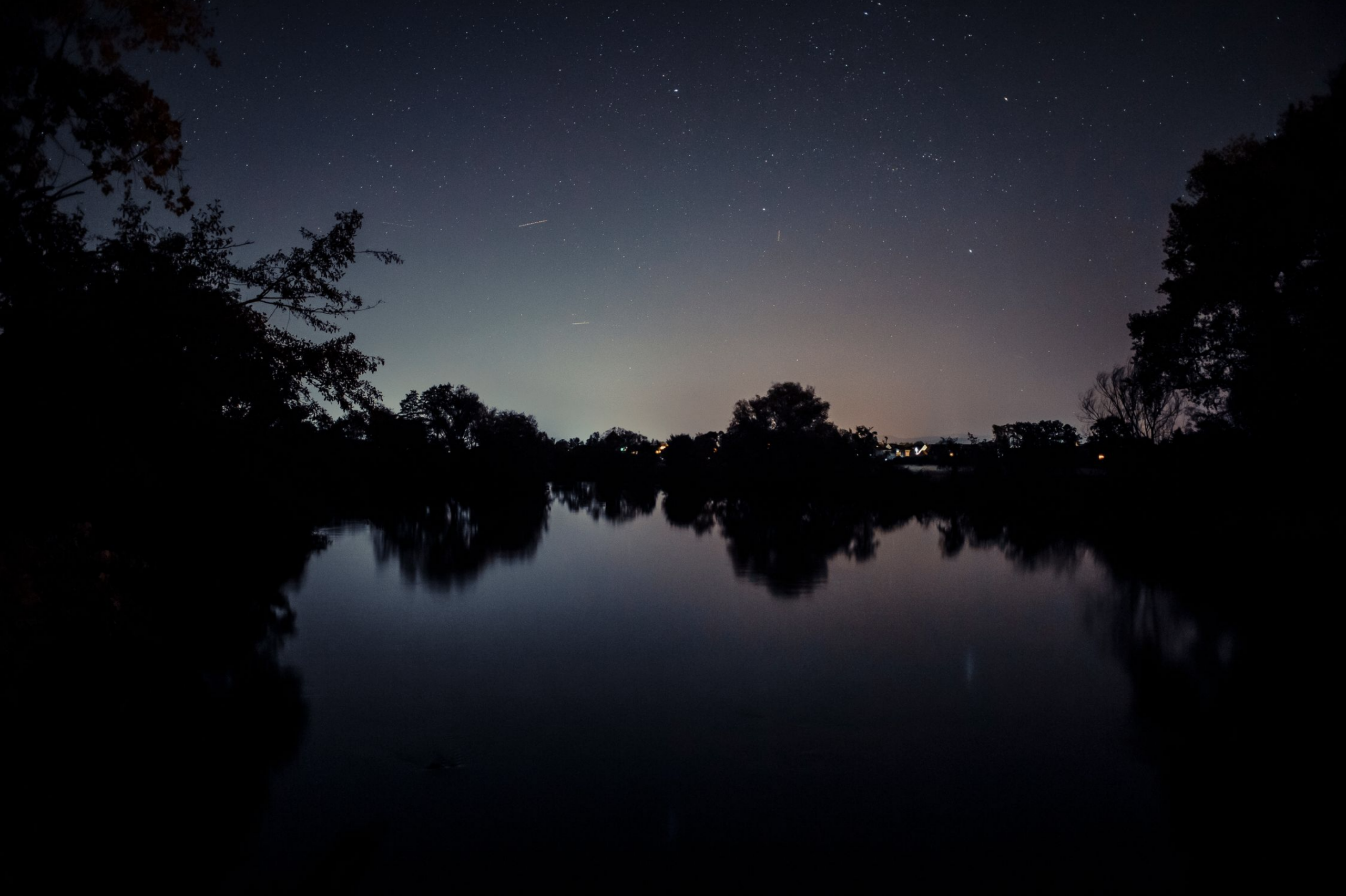 Nachtelijke hemel met sterren boven een stille rivier, omlijst door donkere silhouetten van bomen.