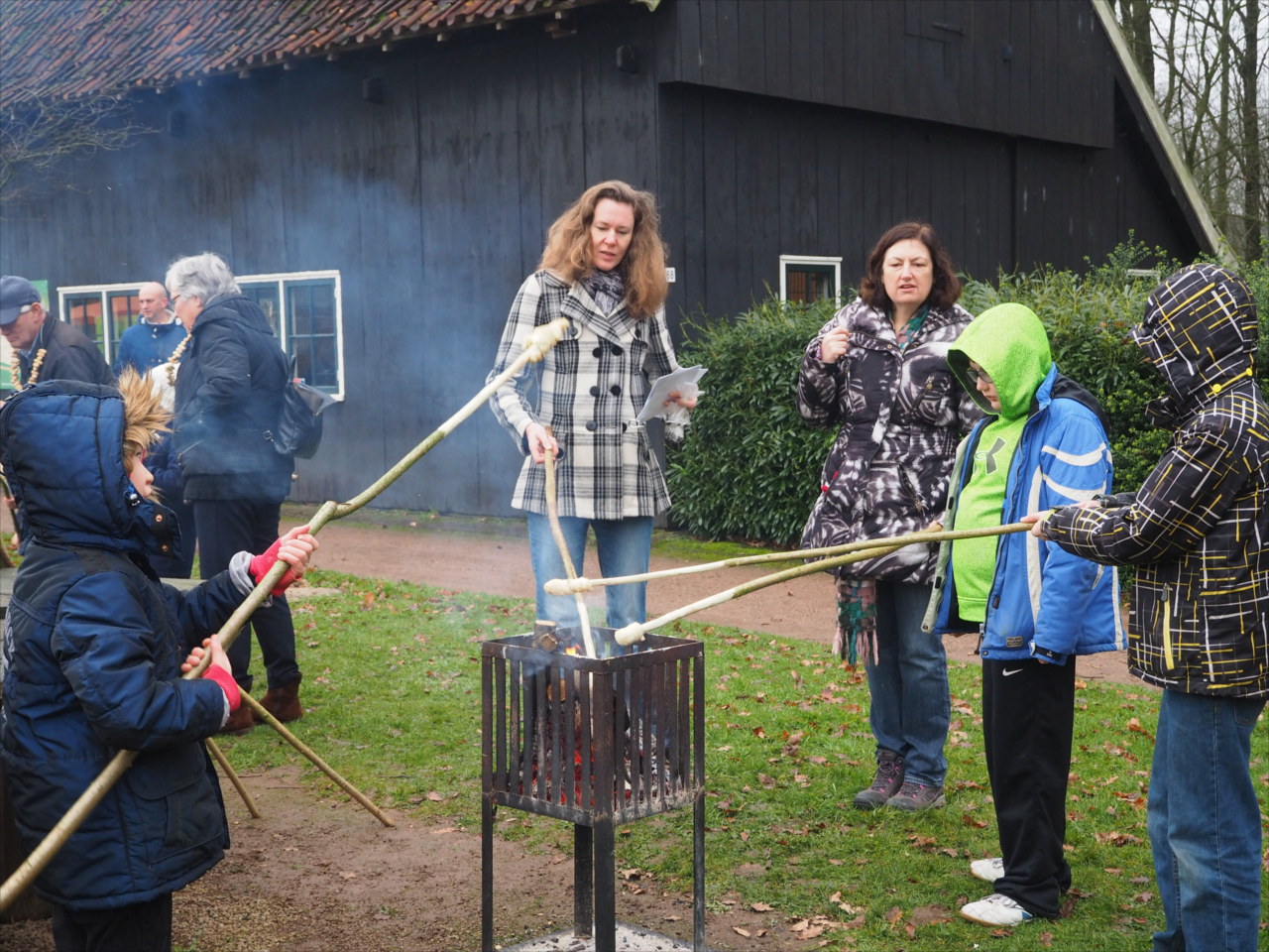 Kinderen roosteren brood op stokken bij een buitenvuur, met volwassenen op de achtergrond.