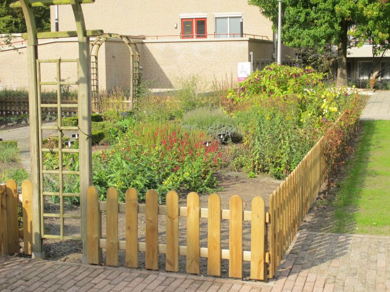 Tuin met houten hek, bloemen en planten, onder een blauwe lucht, naast een gebouw.