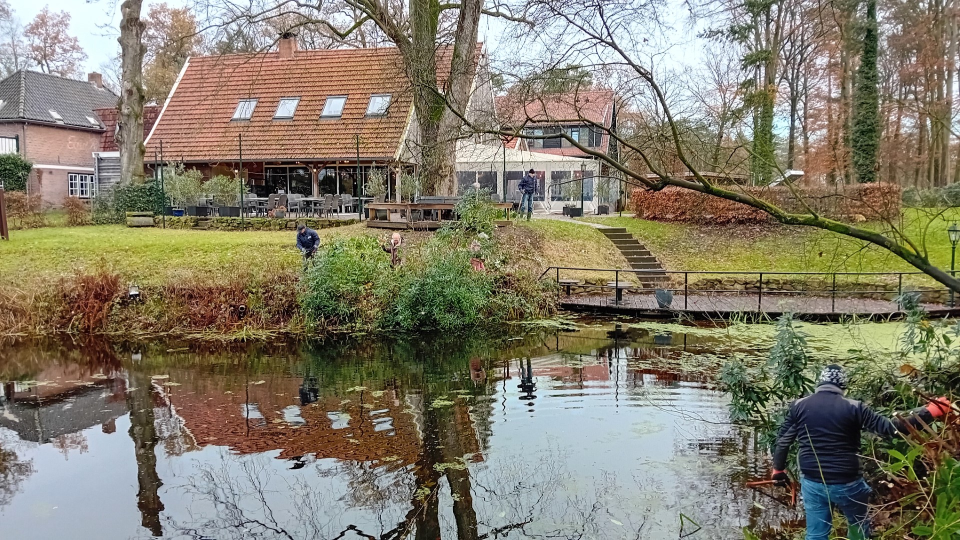 Huis met terras aan een vijver, mensen zijn bezig met tuinwerkzaamheden langs de waterkant.