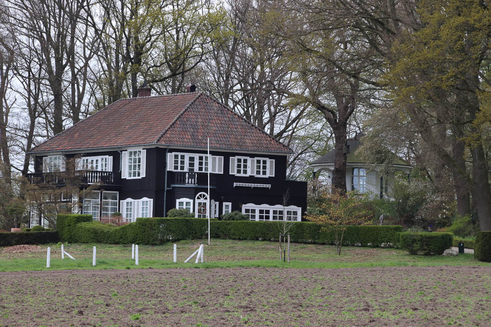 Een groot, zwart huis met witte kozijnen, omgeven door bomen en een heg, naast een veld.
